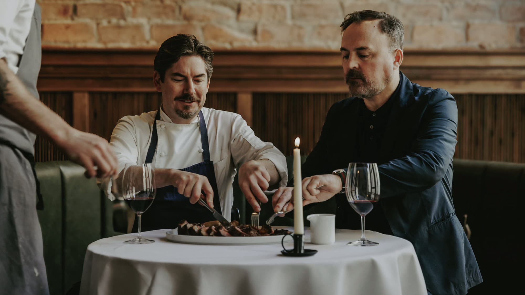 The chef and a guest sharing a plate of sliced steak at a candlelit, white-clothed table at Eleven Fifty Five, brick walls and green leather banquettes behind them